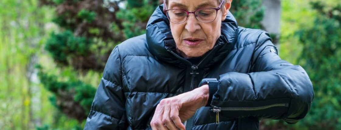 Person im Freien trägt eine dunkle Jacke und blickt auf ein Armband mit integriertem Caritas-Notrufsystem am Handgelenk; im Hintergrund sind Bäume und Grünflächen zu sehen.