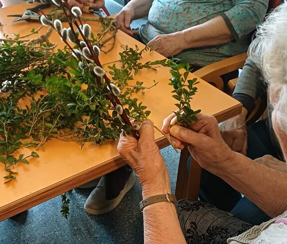 Bewohnerinnen des Pflegewohnhauses gestalten gemeinsam Palmbuschen. Auf dem Tisch liegen frische Zweige und Weidenkätzchen, während sorgfältig gebunden und gearbeitet wird