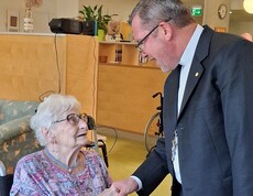 Eine Bewohnerin sitzt an einem Tisch im Aufenthaltsbereich des Caritas Pflegewohnhauses, während Weihbischof Johannes Freitag sie mit einem Handschlag begrüßt und sich mit ihr austauscht.