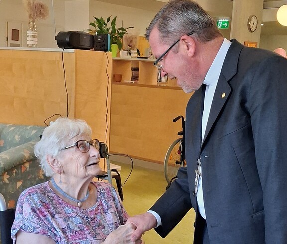 Eine Bewohnerin sitzt an einem Tisch im Aufenthaltsbereich des Caritas Pflegewohnhauses, während Weihbischof Johannes Freitag sie mit einem Handschlag begrüßt und sich mit ihr austauscht.