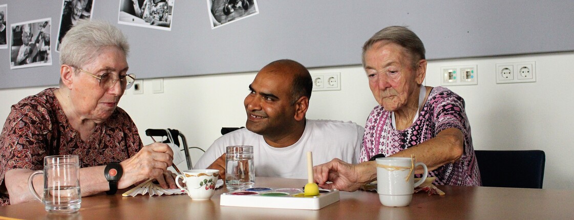 Zwei Seniorinnen sitzen am Tisch und malen, Pfleger in der Mitte