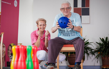 Ein Mann hält eine weiche Bowling-Kugel in den Händen, damit er die Pins im Vordergrund treffen kann. Eine Pflegerin kniet neben ihm und feuert ihn an.