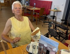 Copilot said:  Eine Bewohnerin sitzt an einem Tisch, hält einen Löffel in der Hand und freut sich auf den Eisbecher vor ihr.