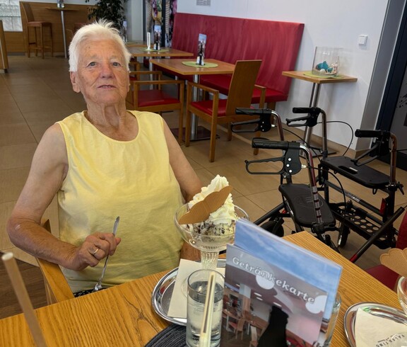 Copilot said:  Eine Bewohnerin sitzt an einem Tisch, hält einen Löffel in der Hand und freut sich auf den Eisbecher vor ihr.