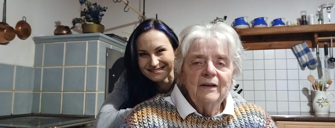 Im Vordergrund ist eine ältere sitzende Dame zu sehen. Dahinter beugt sich eine junge Frau mit langen dunklen Haaren runter und lächelt in die Kamera. 