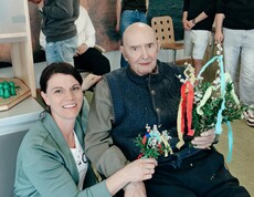 Ein Bewohner des Pflegewohnhauses sitzt im Rollstuhl und hält mehrere bunt geschmückte Palmbuschen in den Händen. Hausleiterin Viktoria Trois kniet unterstützend neben ihm, während im Hintergrund junge Helferinnen und Helfer weitere Zweige vorbereiten.