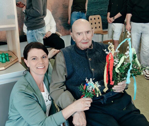 Ein Bewohner des Pflegewohnhauses sitzt im Rollstuhl und hält mehrere bunt geschmückte Palmbuschen in den Händen. Hausleiterin Viktoria Trois kniet unterstützend neben ihm, während im Hintergrund junge Helferinnen und Helfer weitere Zweige vorbereiten.