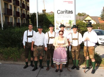 Sechs Mitglieder der Pischelsdorfer Schuhplattler posieren in traditioneller Tracht gemeinsam mit der Pflegedienstleitung vor dem Caritas Pflegewohnhaus Graz-Straßgang im Rahmen des Herbstfestes.