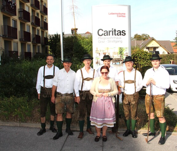 Sechs Mitglieder der Pischelsdorfer Schuhplattler posieren in traditioneller Tracht gemeinsam mit der Pflegedienstleitung vor dem Caritas Pflegewohnhaus Graz-Straßgang im Rahmen des Herbstfestes.