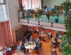 In dem hellen und großzügigen Aufenthaltsbereich eines Pflegewohnhauses sitzen mehrere Bewohnerinnen und Bewohner an Tischen beisammen und genießen das gemeinsame Beisammensein in einer von zahlreichen Pflanzen und herbstlicher Dekoration geprägten Atmosphäre.