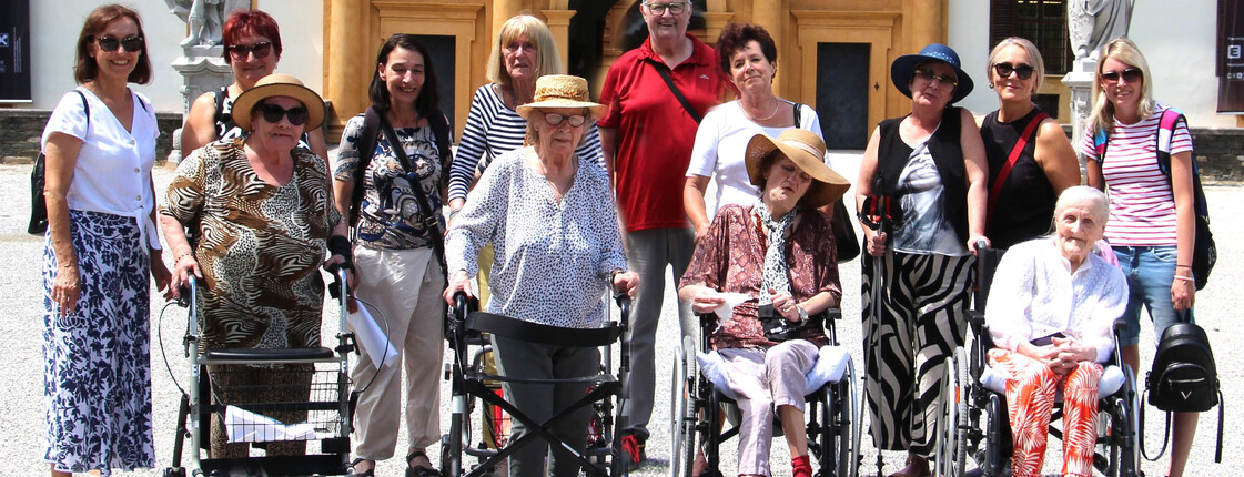 Einige Bewohner*innen sitzen in Rollstühlen bzw. mit Rollatoren stehen nebeneinander. Hinter ihnen befinden sich Pflegepersonal sowie Angehörige. Im Hintergrund ist das Schloss Eggenberg zu sehen.