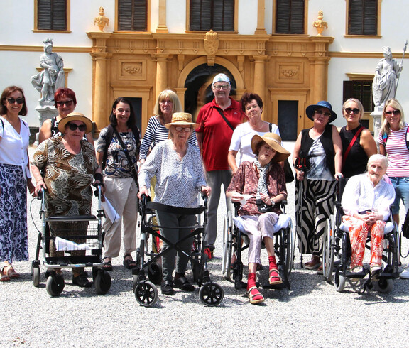 Einige Bewohner*innen sitzen in Rollstühlen bzw. mit Rollatoren stehen nebeneinander. Hinter ihnen befinden sich Pflegepersonal sowie Angehörige. Im Hintergrund ist das Schloss Eggenberg zu sehen.