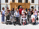 Einige Bewohner*innen sitzen in Rollstühlen bzw. mit Rollatoren stehen nebeneinander. Hinter ihnen befinden sich Pflegepersonal sowie Angehörige. Im Hintergrund ist das Schloss Eggenberg zu sehen.