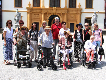 Einige Bewohner*innen sitzen in Rollstühlen bzw. mit Rollatoren stehen nebeneinander. Hinter ihnen befinden sich Pflegepersonal sowie Angehörige. Im Hintergrund ist das Schloss Eggenberg zu sehen.