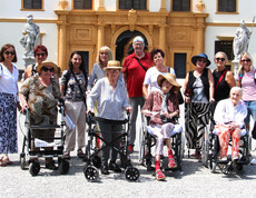 Einige Bewohner*innen sitzen in Rollstühlen bzw. mit Rollatoren stehen nebeneinander. Hinter ihnen befinden sich Pflegepersonal sowie Angehörige. Im Hintergrund ist das Schloss Eggenberg zu sehen.