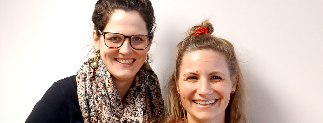 Zwei Frauen im Porträt. Links steht größere Frau mit dunklen Haaren, rechts eine kleinere Frau mit hellen Haaren,