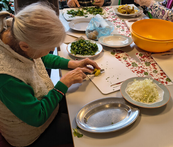 Eine Seniorin schneidet Gemüse, daneben liegen gehackte Kräuter, Zwiebeln und Kartoffeln bereit.