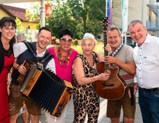 Sechs Personen stehen gemeinsam im Freien. Zwei von ihnen halten Musikinstrumente – eine Person ein Akkordeon, eine andere eine Gitarre. Im Hintergrund hängt ein Banner mit der Aufschrift „20 Jahre“ und „Herzlich Willkommen“. Die Szene wirkt festlich und deutet auf eine Jubiläumsfeier oder ein besonderes Ereignis hin. Von links nach rechts: Claudia Gaube, ein Musiker, Moderatorin Monika Fellner, Jass Gitti, ein Musiker und Josef Url. 
