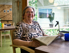 Eine ältere Frau sitzt entspannt an einem Tisch in einem Gemeinschaftsraum. Sie hält eine Zeitung in den Händen und liest darin. Auf dem Tisch vor ihr stehen eine blaue Tasse und weitere Gegenstände. Hinter ihr befindet sich eine große Fensterfront mit Blick auf Grünpflanzen sowie ein Rollator.