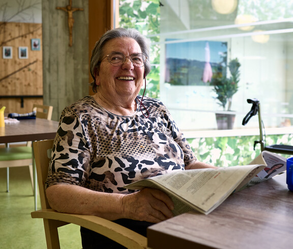 Eine ältere Frau sitzt entspannt an einem Tisch in einem Gemeinschaftsraum. Sie hält eine Zeitung in den Händen und liest darin. Auf dem Tisch vor ihr stehen eine blaue Tasse und weitere Gegenstände. Hinter ihr befindet sich eine große Fensterfront mit Blick auf Grünpflanzen sowie ein Rollator.