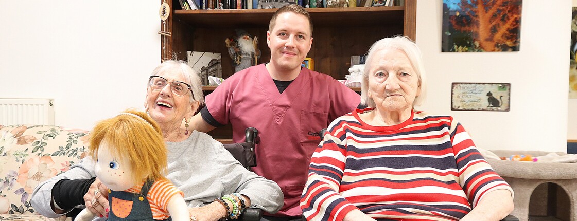 Junger Mann steht hinter zwei sitzenden älteren Damen.