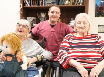 Junger Mann steht hinter zwei sitzenden älteren Damen.