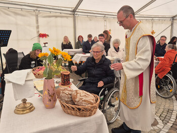 Eine Person in religiösen Gewändern interagiert mit einer älteren Person im Rollstuhl. Der Tisch ist mit Sonnenblumen und Brot dekoriert. Mehrere Personen sitzen an Tischen im Zelt.
