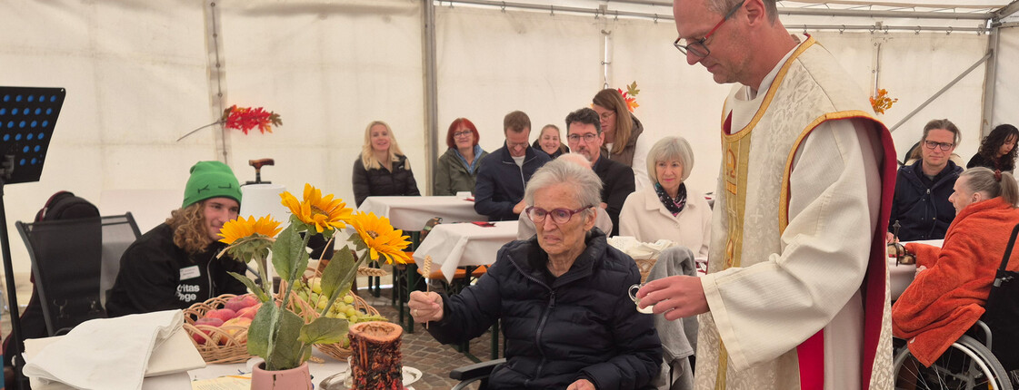 Eine Person in religiösen Gewändern interagiert mit einer älteren Person im Rollstuhl. Der Tisch ist mit Sonnenblumen und Brot dekoriert. Mehrere Personen sitzen an Tischen im Zelt.