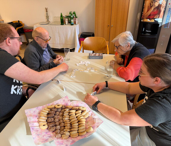 Bewohner*innen und Jugendliche sitzen am Tisch und legen gemeinsam Dominosteine im Festsaal aus.