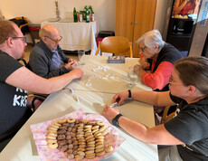 Bewohner*innen und Jugendliche sitzen am Tisch und legen gemeinsam Dominosteine im Festsaal aus.