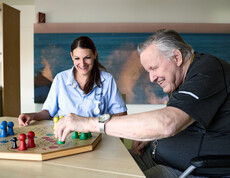 Eine Pflegekraft und eine ältere Person sitzen gemeinsam an einem Tisch und spielen ein Brettspiel mit bunten Spielfiguren. Die Pflegekraft trägt Dienstkleidung und unterstützt die Person, die im Rollstuhl sitzt. Im Hintergrund ist eine ruhige Wanddekoration mit Meeresmotiv zu sehen.