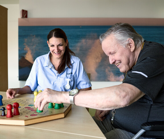 Eine Pflegekraft und eine ältere Person sitzen gemeinsam an einem Tisch und spielen ein Brettspiel mit bunten Spielfiguren. Die Pflegekraft trägt Dienstkleidung und unterstützt die Person, die im Rollstuhl sitzt. Im Hintergrund ist eine ruhige Wanddekoration mit Meeresmotiv zu sehen.