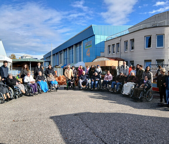 Große Gruppe von Menschen, viele in Rollstühlen, vor einem Gebäudekomplex bei sonnigem Wetter.