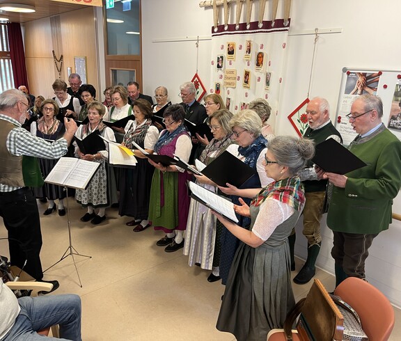 Ein Chor trägt Lieder im Gemeinschaftsraum eines Pflegewohnhauses vor, die Sängerinnen und Sänger halten Notenmappen