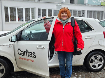 Eine Frau mit roter Jacke steht vor einem Caritas Auto mit offener Fahrertür