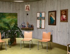 Ein runder Raum mit Betonwänden ist mit mehreren religiösen Bildern dekoriert, darunter ein Mosaikkreuz und Darstellungen von Heiligen. Vor der Wand stehen drei helle Holzstühle in einer Halbkreis-Anordnung. Links befindet sich eine große grüne Zimmerpflanze, daneben ein kleines Wandregal mit einer Marienfigur, einem Kreuz und Kerzen.