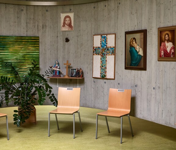 Ein runder Raum mit Betonwänden ist mit mehreren religiösen Bildern dekoriert, darunter ein Mosaikkreuz und Darstellungen von Heiligen. Vor der Wand stehen drei helle Holzstühle in einer Halbkreis-Anordnung. Links befindet sich eine große grüne Zimmerpflanze, daneben ein kleines Wandregal mit einer Marienfigur, einem Kreuz und Kerzen.