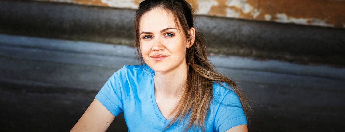 Eine junge Frau mit braunen langen Haaren und Pferdeschwanz sitzt auf einer Stiege, sie trägt ein hellblaues T-Shirt mit Caritas Logo und lächelt in die Kamera.