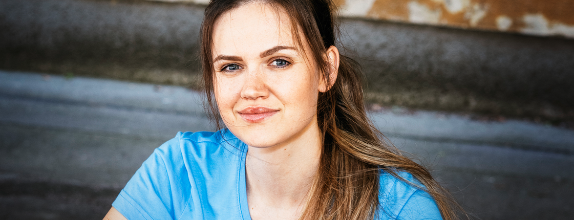 Caritas Pflege: Wir sind die Antwort auf Who Cares. Eine junge Frau mit braunen langen Haaren und Pferdeschwanz sitzt auf einer Stiege, sie trägt ein hellblaues T-Shirt mit Caritas Logo und lächelt in die Kamera.