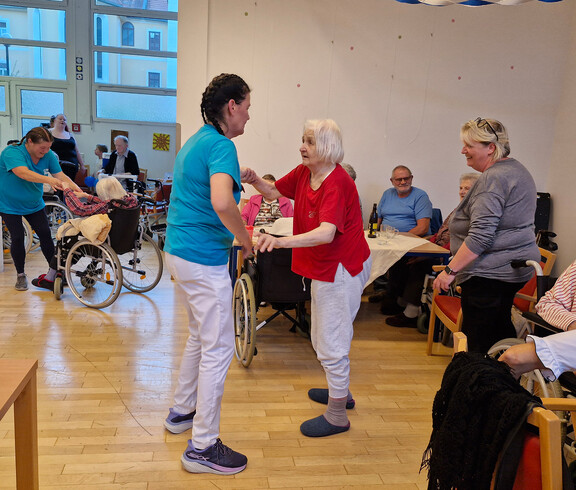 Senior*innen tanzen mit Pflegekräften, andere Gäste schauen lächelnd zu