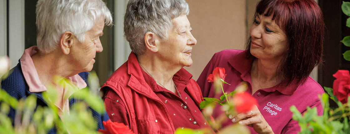 Zwei ältere Damen stehen mit einer Pflegekraft am Balkon und unterhalten sich. Vor ihnen sieht man die Blumen, die in den Blumenkästen wachsen.