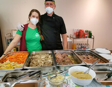 Zwei Personen, die hinter einem Buffet mit verschiedenen Speisen stehen. Beide tragen Gesichtsmasken und Schutzhandschuhe. Die Frau trägt ein grünes T-Shirt der Caritas und der Mann ist in schwarzer Kochkleidung. Vor ihnen sind Tabletts mit Gemüse, Fleischgerichten und Saucen aufgestellt. 