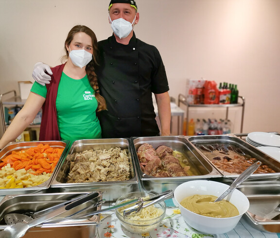 Zwei Personen, die hinter einem Buffet mit verschiedenen Speisen stehen. Beide tragen Gesichtsmasken und Schutzhandschuhe. Die Frau trägt ein grünes T-Shirt der Caritas und der Mann ist in schwarzer Kochkleidung. Vor ihnen sind Tabletts mit Gemüse, Fleischgerichten und Saucen aufgestellt. 