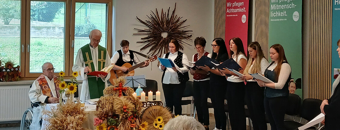 Zwei Geistliche feiern den Erntedank-Gottesdienst vor einer musizierenden Gruppe und herbstlichen Erntegaben.