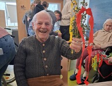 Ein Bewohner des Pflegewohnhauses präsentiert stolz ihren selbstgebundenen Palmbuschen, der mit bunten Bändern verziert ist. Auf dem Tisch liegen Zweige und Bastelmaterial, während im Hintergrund weitere Bewohnerinnen und Bewohner gemeinsam an ihren Palmbuschen arbeiten.