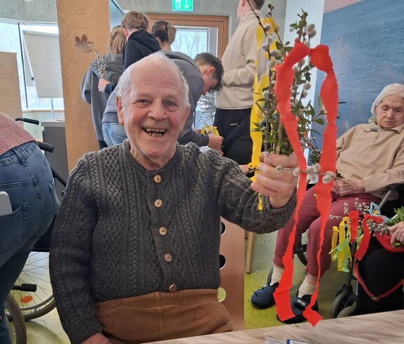 Ein Bewohner des Pflegewohnhauses präsentiert stolz ihren selbstgebundenen Palmbuschen, der mit bunten Bändern verziert ist. Auf dem Tisch liegen Zweige und Bastelmaterial, während im Hintergrund weitere Bewohnerinnen und Bewohner gemeinsam an ihren Palmbuschen arbeiten.