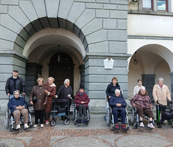 Menschen, einige in Rollstühlen, vor einem Gebäude mit gewölbten Türen und Steinfassade.