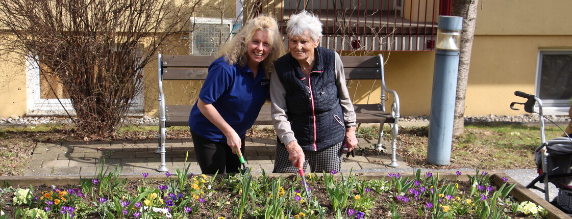 Eine Bewohnerin und eine Mitarbeiterin stehen gemeinsam an einem erhöhten Blumenbeet im Freien und arbeiten mit kleinen Gartengeräten in der Erde. Vor ihnen blühen zahlreiche bunte Frühblüher wie Stiefmütterchen und Krokusse. Im Hintergrund befinden sich eine Bank, ein Baum sowie das Pflegewohnhaus. Ein Rollator steht am rechten Bildrand.