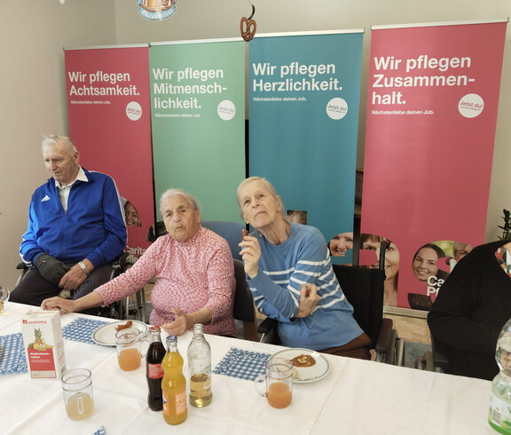 Gruppe älterer Menschen sitzt an gedecktem Tisch, im Hintergrund Caritas-Pflege-Banner.