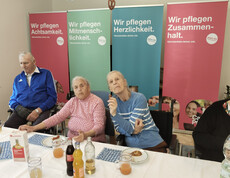 Gruppe älterer Menschen sitzt an gedecktem Tisch, im Hintergrund Caritas-Pflege-Banner.